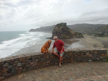 Mature man sitting with dog on retaining wall against sea at beach