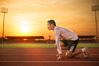 Side view of teenage boy kneeling on sports track against sky during sunset