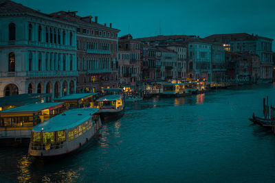 Boats moored in illuminated canal by buildings against sky at dusk