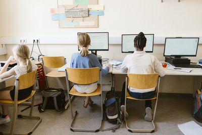 Rear view of multiracial female students using computers while sitting on chairs in classroom at school