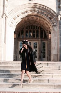 Full length of young woman in graduation gown moving down on steps against college