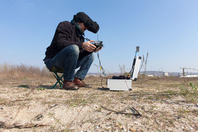 Man operating drone with remote control while sitting on field against sky