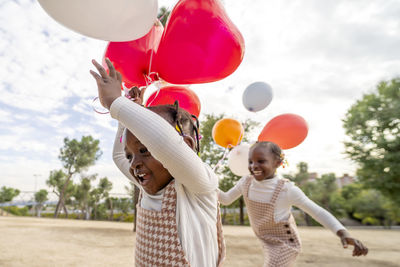 Happy african american little sisters in similar dresses standing with colorful balloons in hands on green grass in park in daylight