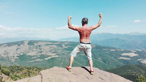 Rear view of shirtless man flexing muscles while standing on cliff against sky