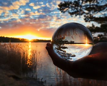 Reflection of person hand holding crystal ball against sky during sunset