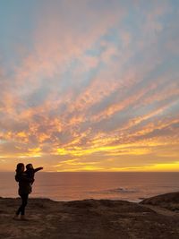 Silhouette mother carrying boy while standing on beach against sky during sunset