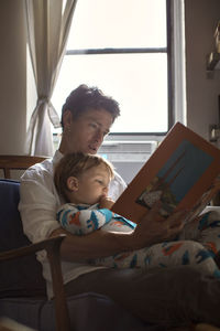 Father reading picture book for son while sitting on chair against window