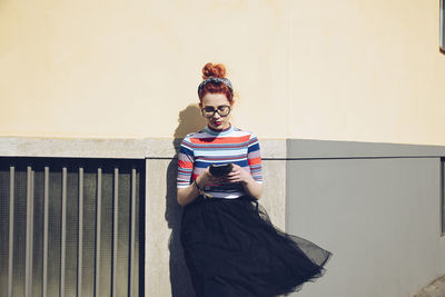 Young female hipster using mobile phone while standing against wall