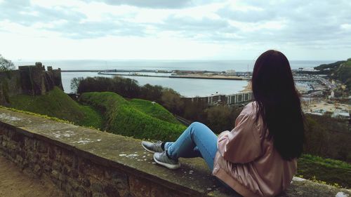 Woman sitting on retaining wall in city against sky