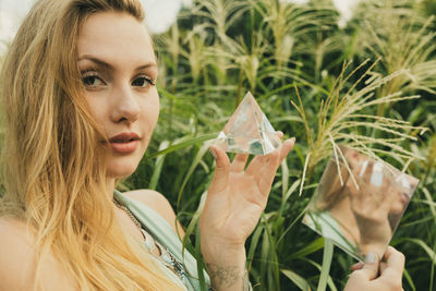 Portrait of young woman holding prism and mirror while standing against plant