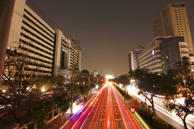 Light trails on city street by buildings against sky at night