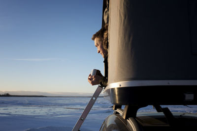 Man looking away while sitting in roof tent on car against sky during sunset