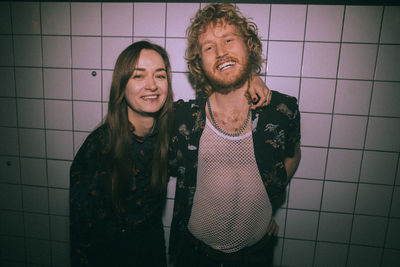 Portrait of young woman standing with arm around bearded male friend against white wall at nightclub
