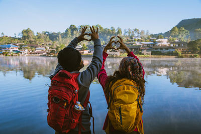 Rear view of people making heart shape with hands while standing by lake against sky