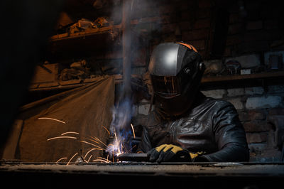 Young mechanic with a welding machine in an old dirty garage at night. man in protection doing