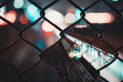 Train at railroad station platform in illuminated city seen through chainlink fence at night