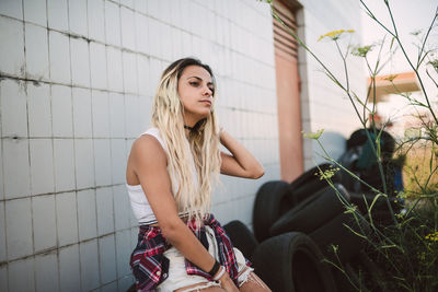 Young woman looking away while sitting on tire against tiled wall