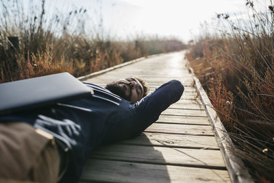 Man with laptop lying down amidst field on boardwalk during sunny day