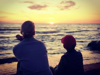 Rear view of people sitting at beach against sky during sunset