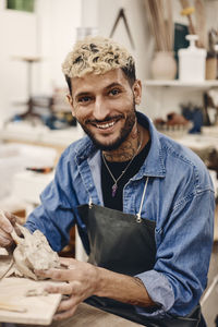Portrait of happy young man molding clay while sitting at table in art class
