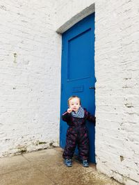 Cute baby boy, barely a toddler, standing alone outside a blue wooden front door chewing a straw