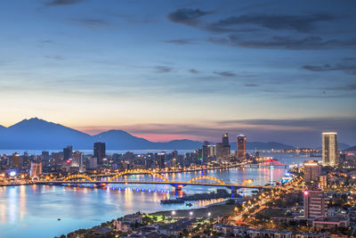 Illuminated city by river against sky during sunset