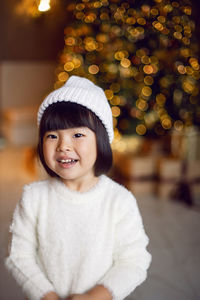 Beautiful korean child girl in in a white knitted sweater and hat stands at the christmas tree 