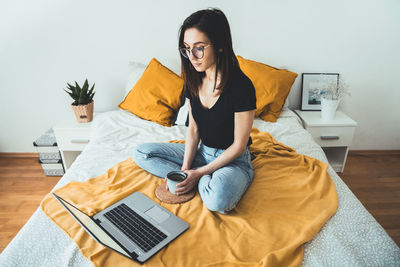 Young woman relaxing and holding cup of hot coffee. female sitting on bed and surfing the net