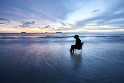 Silhouette man sitting on chair at beach against sky during sunset