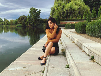 Portrait of young woman sitting by lake against trees