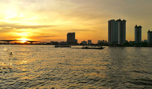 River by cityscape against sky during sunset