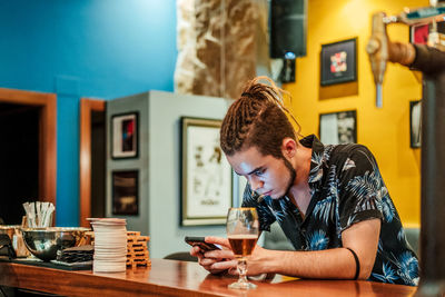 Male bartender leaning on wooden counter and chatting on social media via cellphone while having break during work in bar