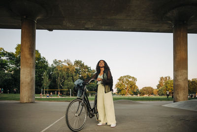 Teenage girl with bicycle standing below bridge