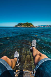 Low section of man on rock by sea against clear blue sky