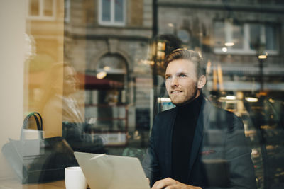 Smiling businessman with laptop looking away in cafe seen through glass window