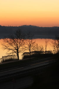 Silhouette bare trees by lake against sky during sunset