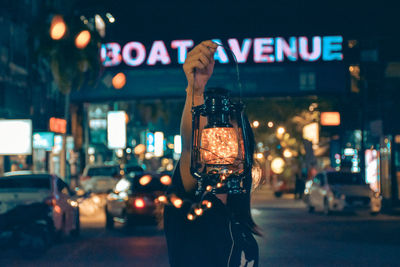Woman holding illuminated lantern in front of face while standing in city at night