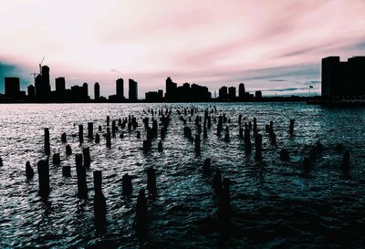 Silhouette wooden posts in sea by city against sky during sunset