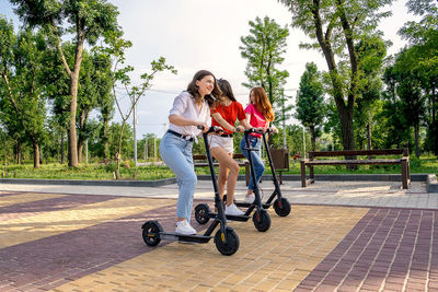 Three young girl friends on vacation having fun driving electric scooter through the city park