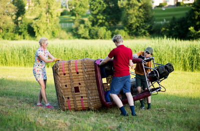 Boys assisting pilot in preparing hot air balloon on grassy field during sunset