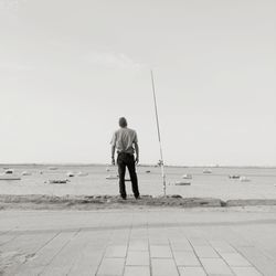 Rear view of man standing by fishing rod at beach against clear sky
