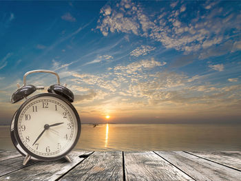 Clock on table by sea against sky during sunset