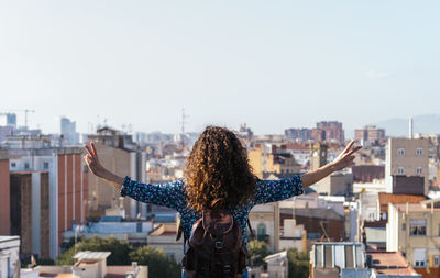 Back view of female with curly hair standing with outstretched arms on roof and showing two fingers while admiring amazing cityscape