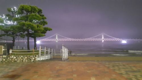 Illuminated suspension bridge over sea against sky