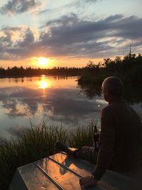 Man sitting with drinks by lake against sky during sunset
