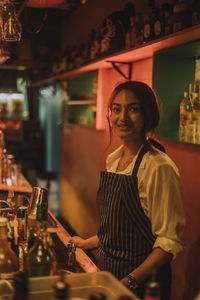 Portrait of smiling female bartender wearing apron and standing at bar
