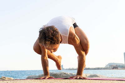 Ground level of middle aged male doing bakasana pose against cloudless blue sky during yoga session on beach in summer