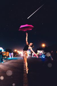 Ballet dancer holding pink umbrella while standing on street against sky at night