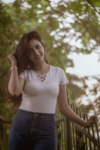 Portrait of smiling woman standing by railing against tree