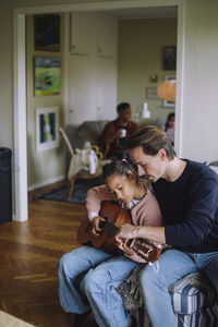 Father sitting with daughter playing guitar in bedroom at home